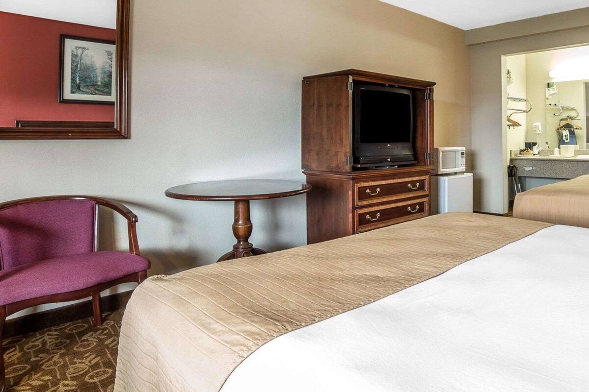 Habitación de hotel con cama, un mueble bajo para televisión de madera, una mesita redonda pequeña, una silla y un espejo de baño.