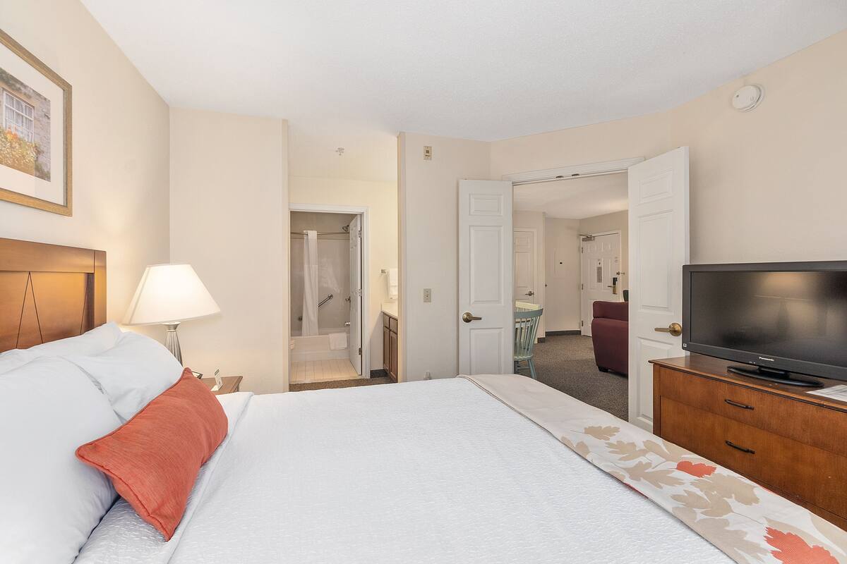 A hotel room with a bed, a TV on a wooden dresser, a bathroom with a shower, and a door leading to another room.