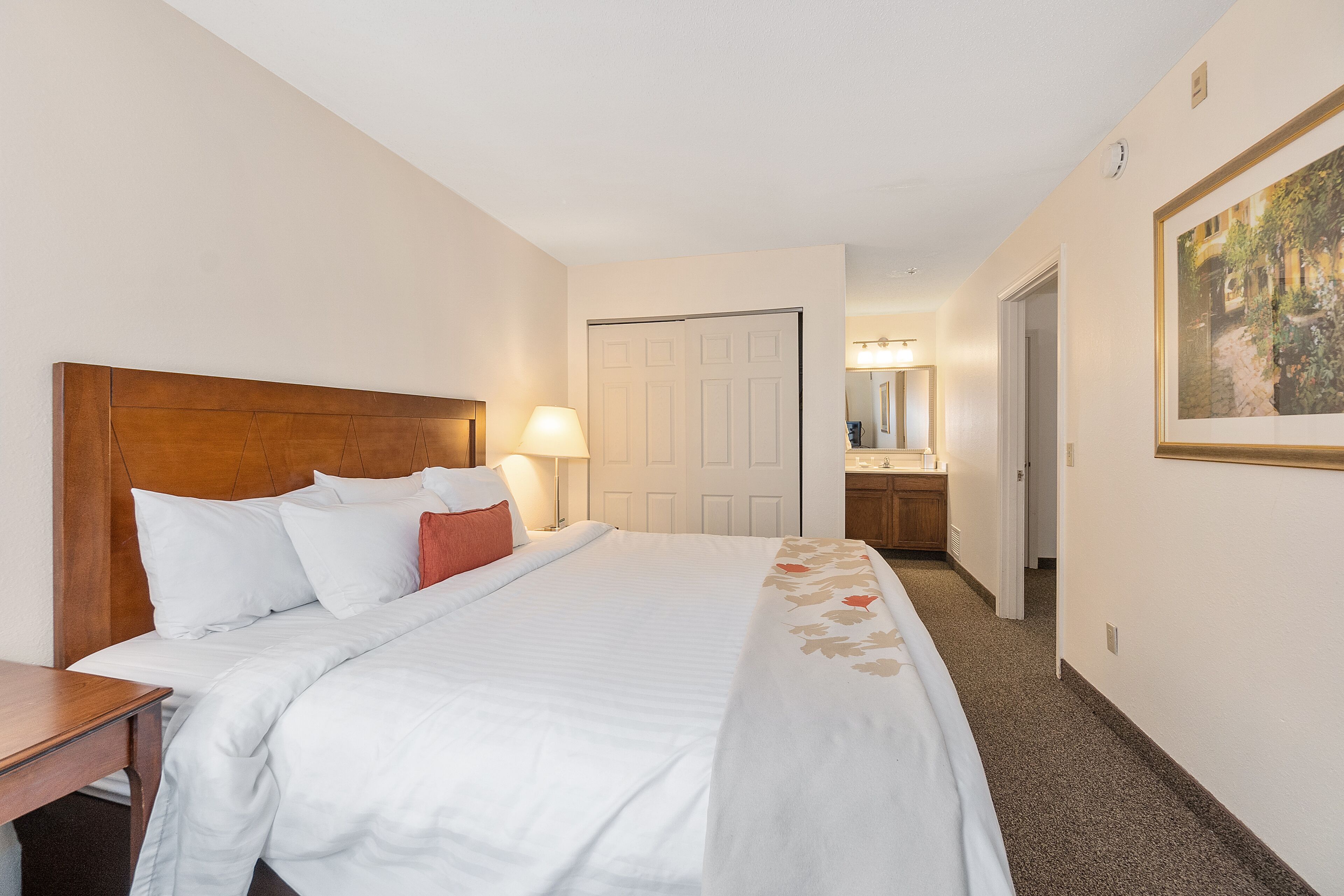 Habitación de hotel con una cama grande, cabecera de madera, mesita de noche, lámpara, puerta, baño con espejo y un cuadro enmarcado en la pared.