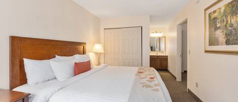 Habitación de hotel con una cama grande, cabecera de madera, mesita de noche, lámpara, puerta, baño con espejo y un cuadro enmarcado en la pared.