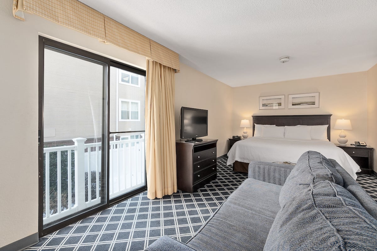 A hotel room with a large sliding glass door to the balcony, a bed with white linens, a TV on a dresser, and a gray sofa.