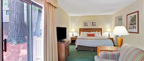 A hotel room with a large sliding glass door, a bed with two pillows, a bedside table with a lamp, a striped sofa, and a television on a wooden dresser.
