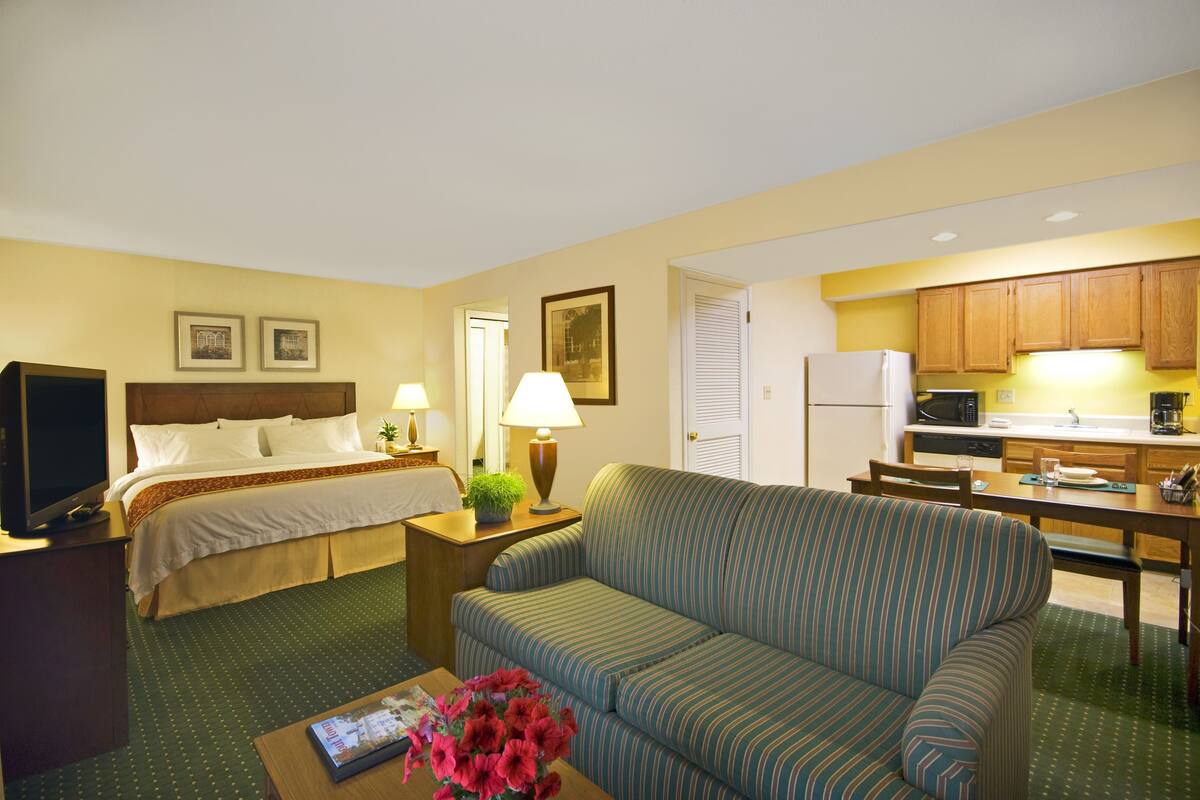 A hotel room with a bed, a sofa, a dining table, and a kitchen area.