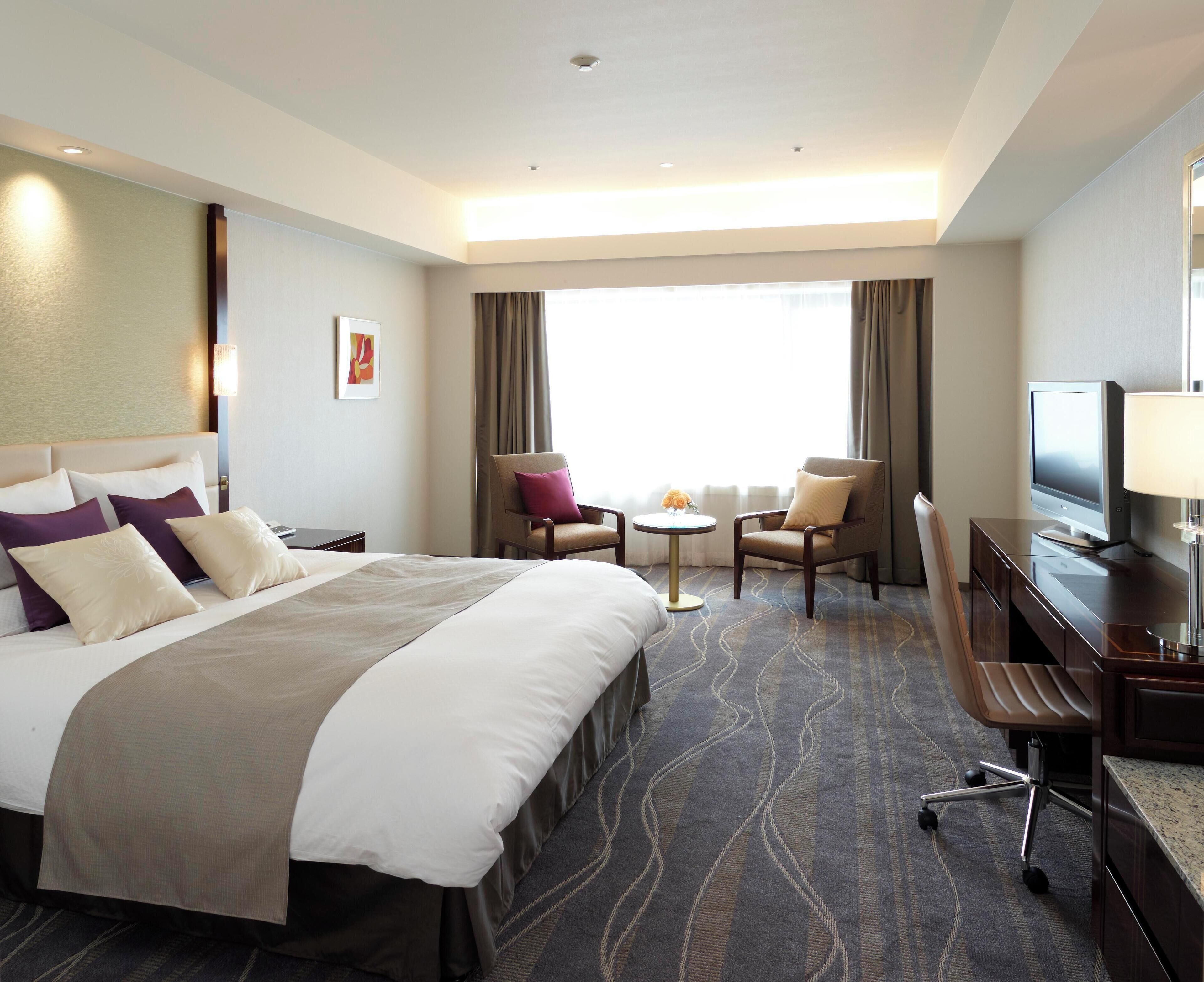 A hotel room with a large bed, a desk, a chair, a television, and a small table with flowers.
