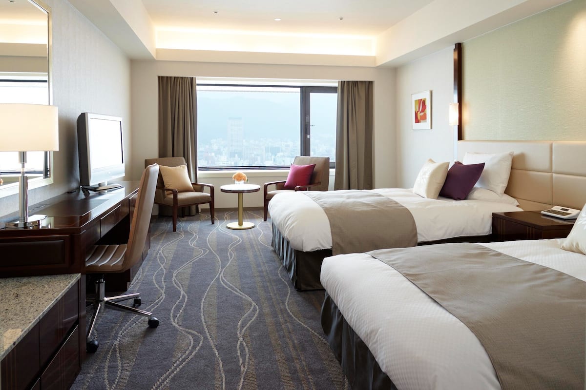 A hotel room with two beds, a desk, a chair, a small table, and a view of the city.