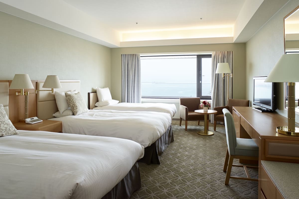 A hotel room with two beds, a desk, a chair, a TV, and a view of the sea.