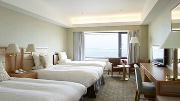 A hotel room with two beds, a desk, a chair, a TV, and a view of the sea.