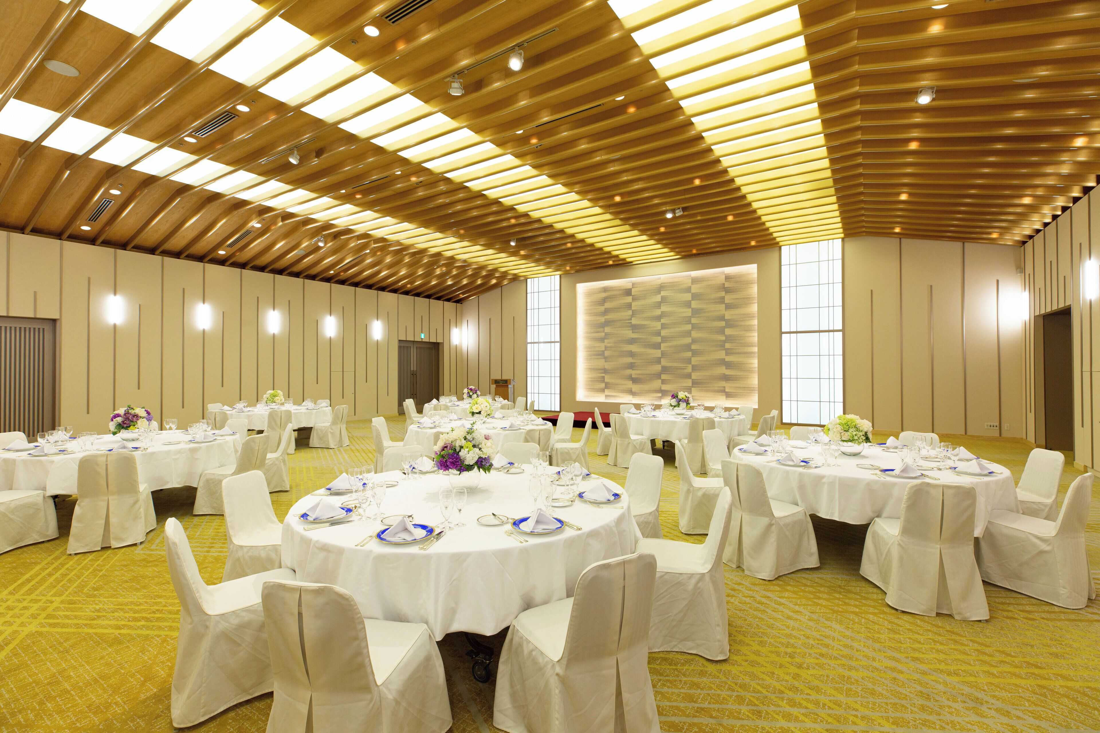 A banquet hall with multiple round tables set for dining, white chairs, and a high ceiling with wooden paneling.