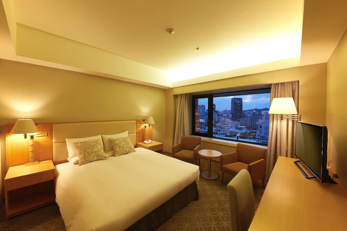 A hotel room with a bed, bedside tables, a desk, a chair, a TV, and a window with a city view.