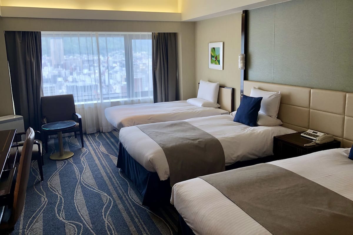 A hotel room with two beds, a desk, a chair, and a large window with a city view.
