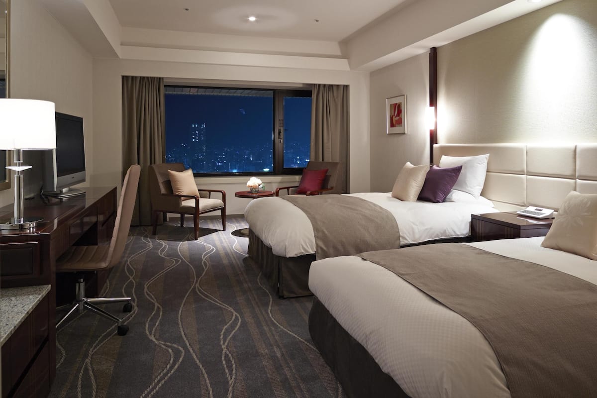 A hotel room with two beds, a desk with a computer, a chair, a window with a city view, and a nightstand with a lamp.
