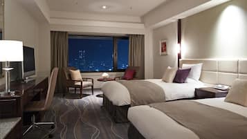 A hotel room with two beds, a desk with a computer, a chair, a window with a city view, and a nightstand with a lamp.