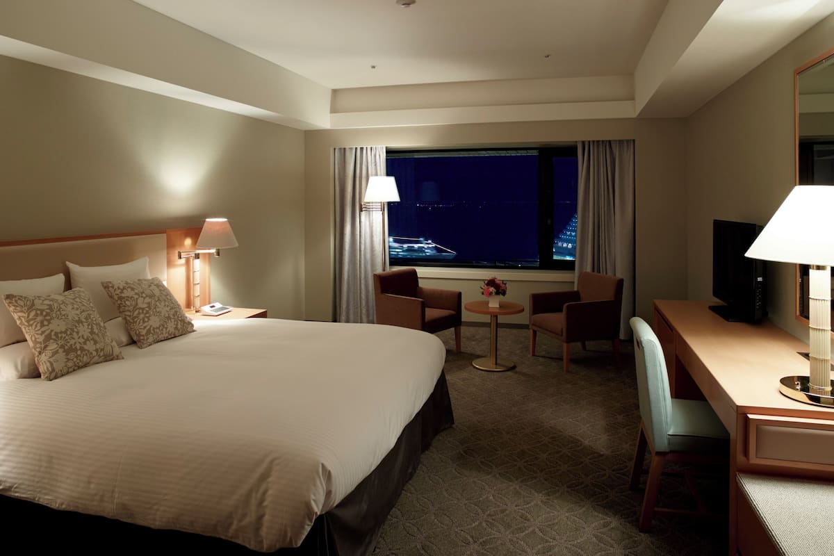 A hotel room with a large bed, a desk with a chair, a television, and a window with a night view of a cityscape.