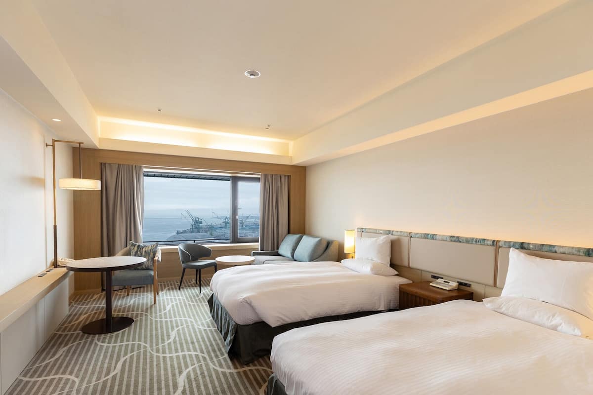 A hotel room with two beds, a small round table, a chair, and a view of the sea.