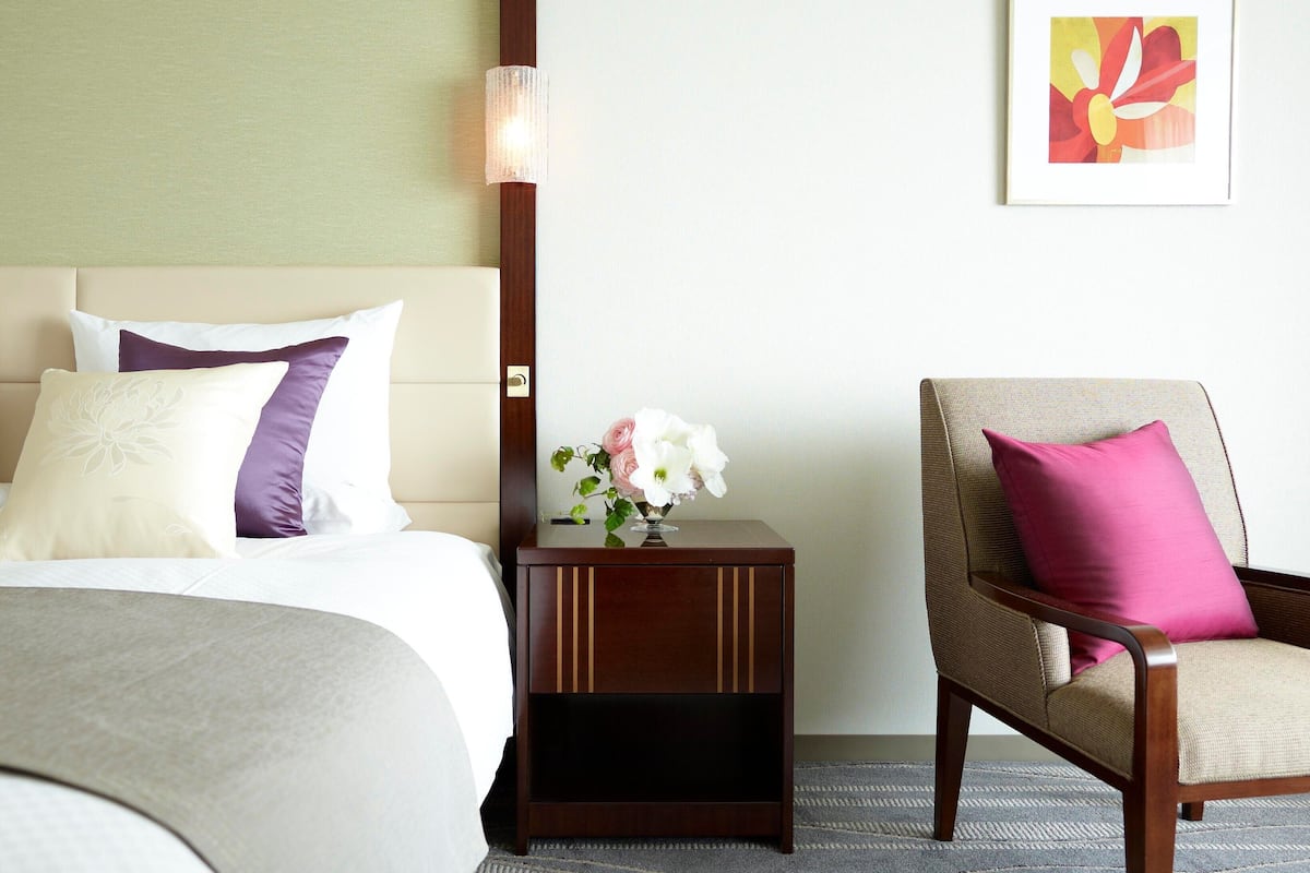 A hotel room with a bed, a nightstand with a vase of flowers, a chair with a cushion, and a framed picture on the wall.