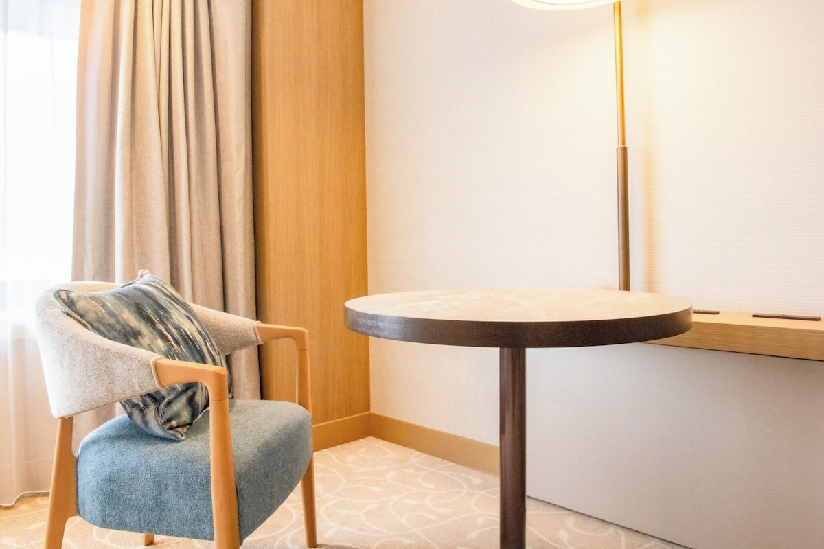 A corner of a hotel room with a round table, a chair with a cushion, a lamp, and a curtain.