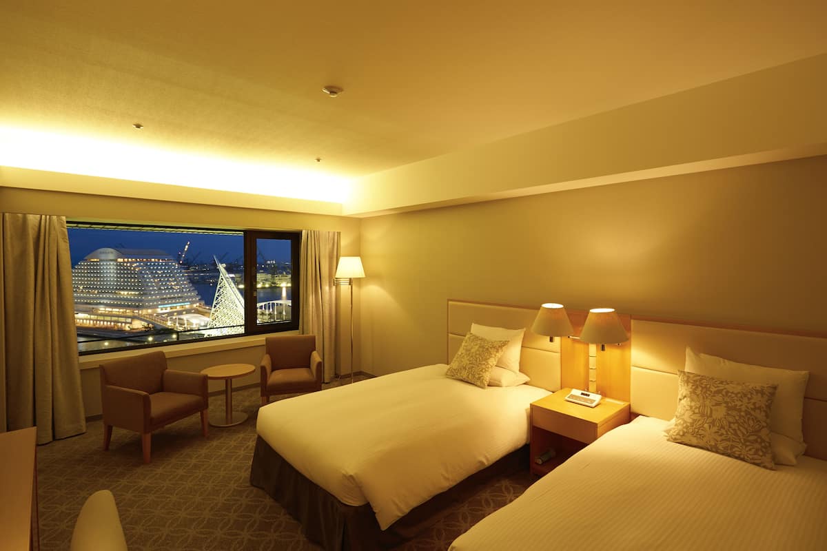 A hotel room with two beds, a small table, a chair, and a view of a cityscape at night.