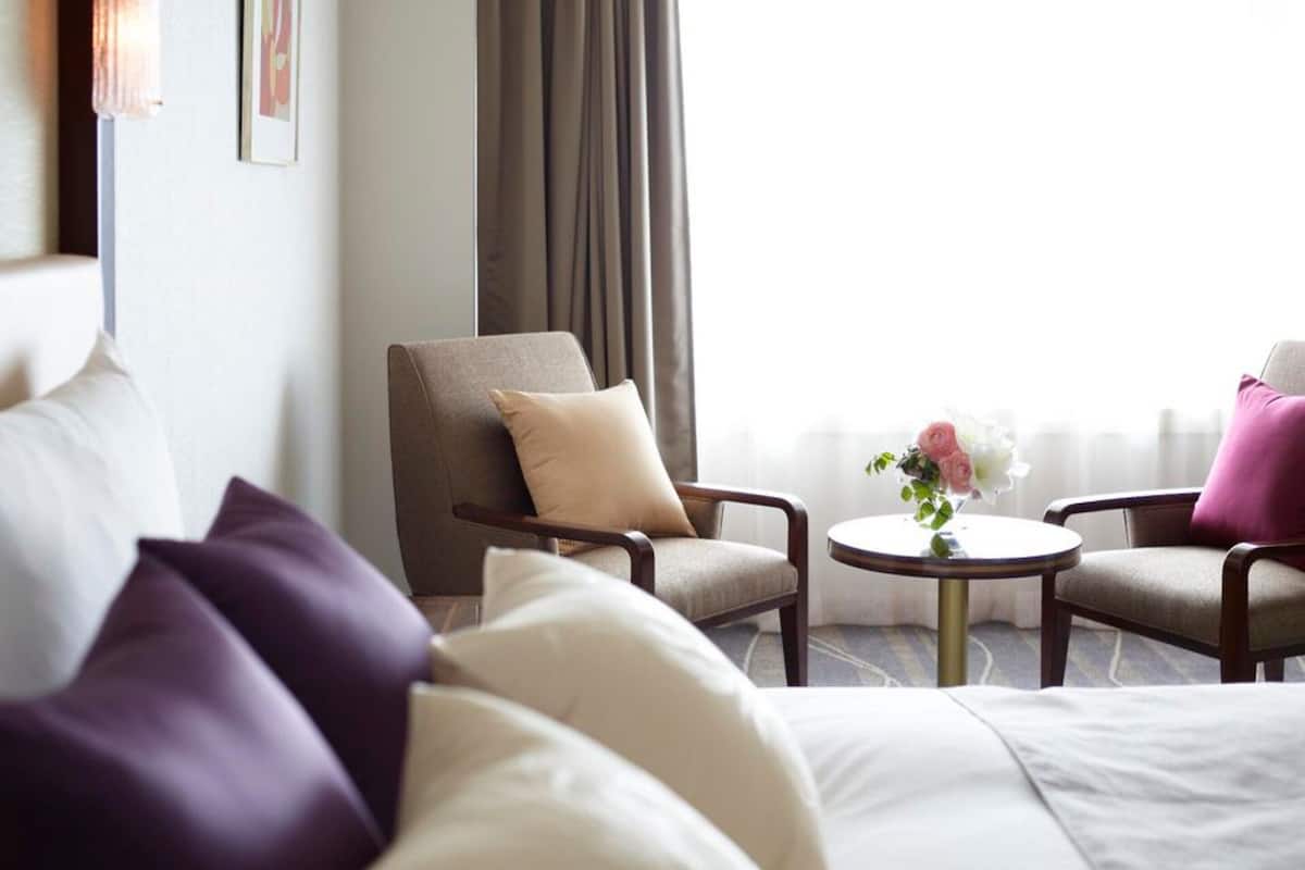 A hotel room with a bed, two armchairs, a small round table with a vase of flowers, and a window with curtains.