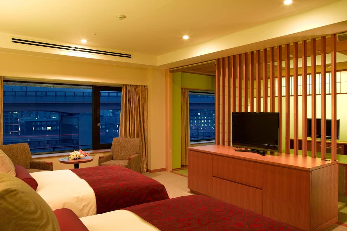 A hotel room with two beds, a TV, a desk, and large windows with a city view.