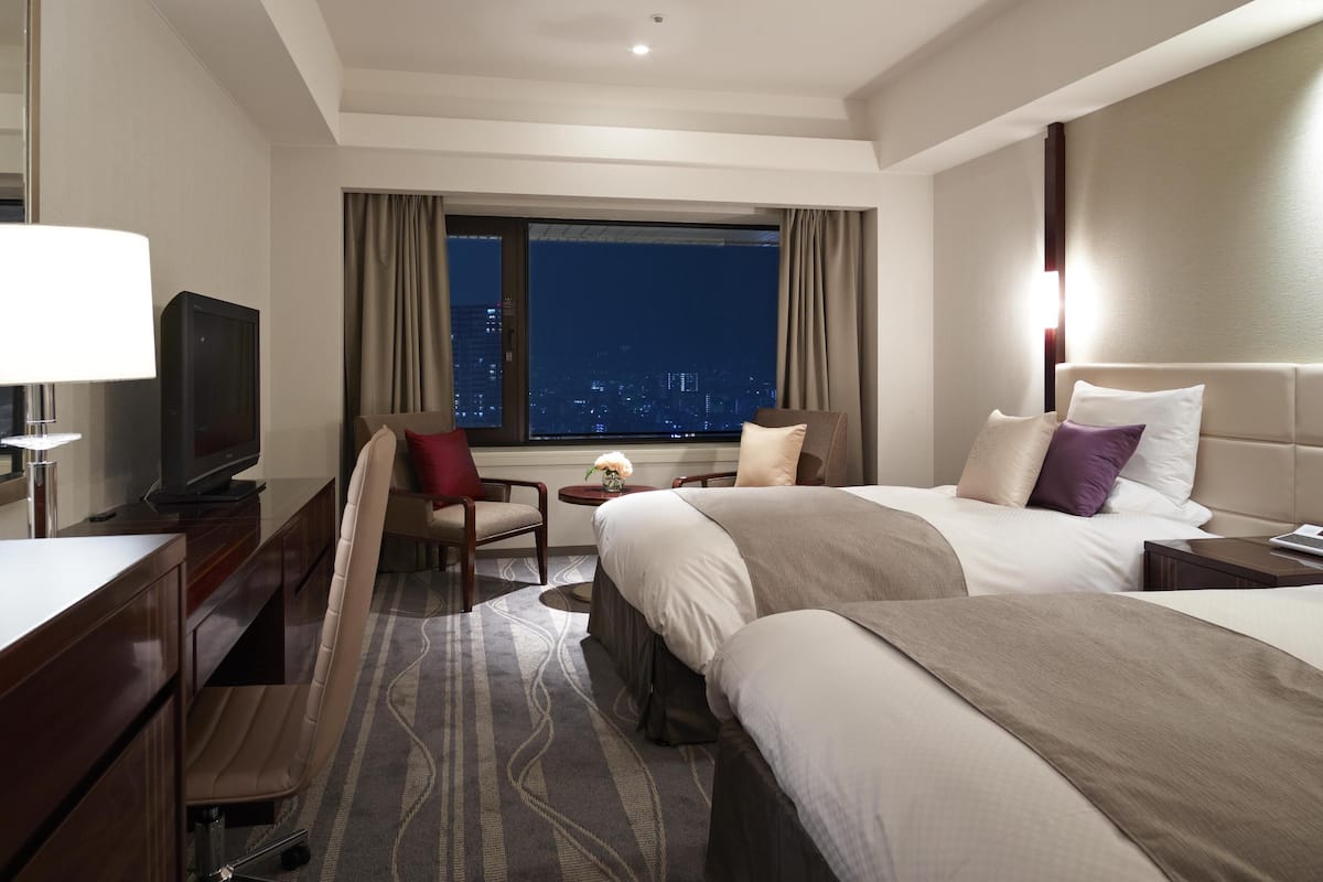 A hotel room with a large bed, a desk with a lamp, a chair, and a TV. There is a window with a city view.