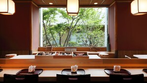 A restaurant with wooden tables and benches, a view of trees through a large window, and hanging pendant lights. - Hotel Okura Kobe (Kobe)