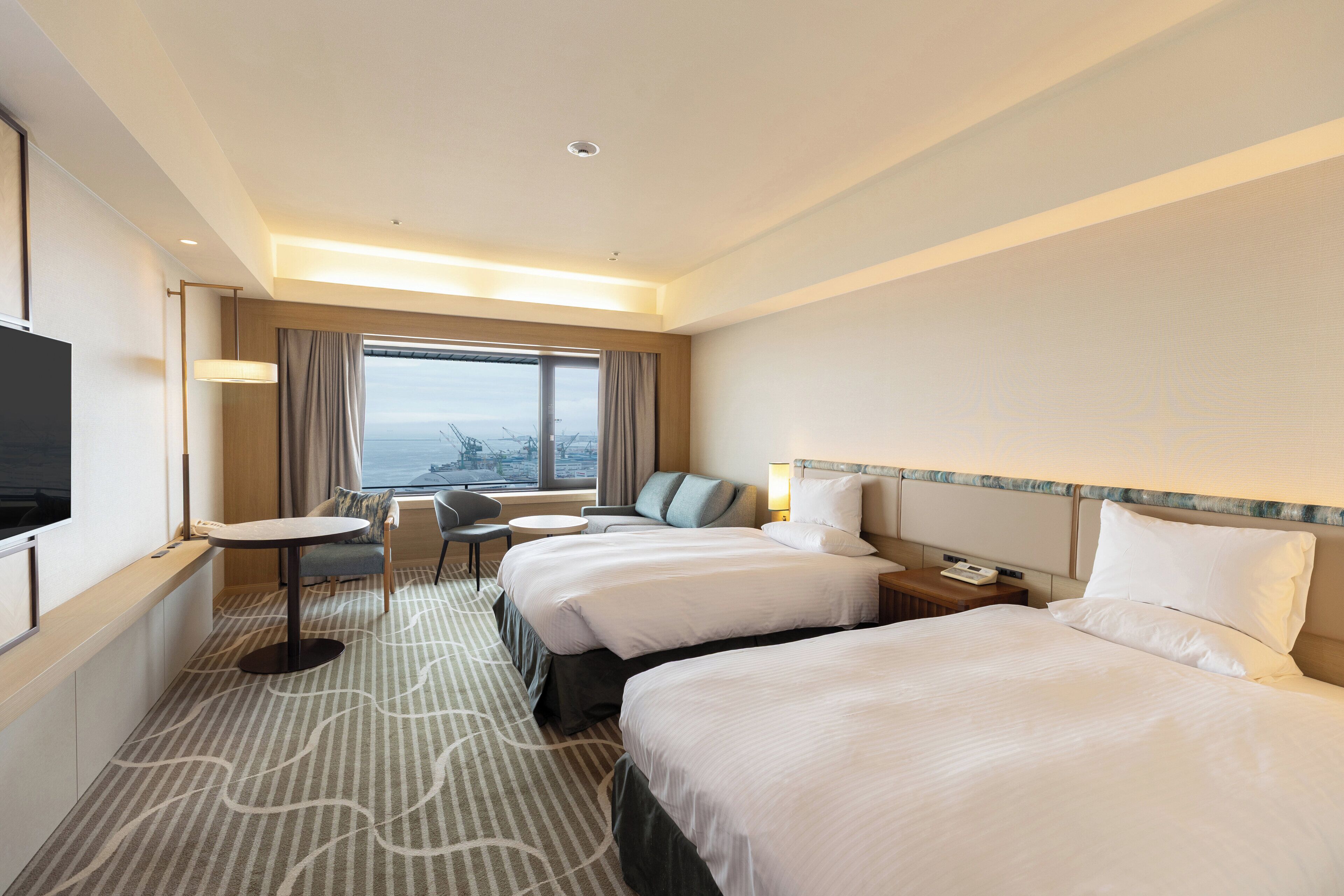 A hotel room with two beds, a desk, a chair, a TV, and a window with a city view.