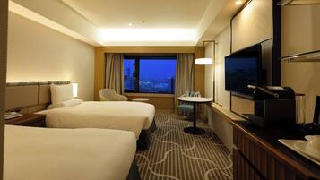 A hotel room with two beds, a TV, a desk, and a window with a city view.