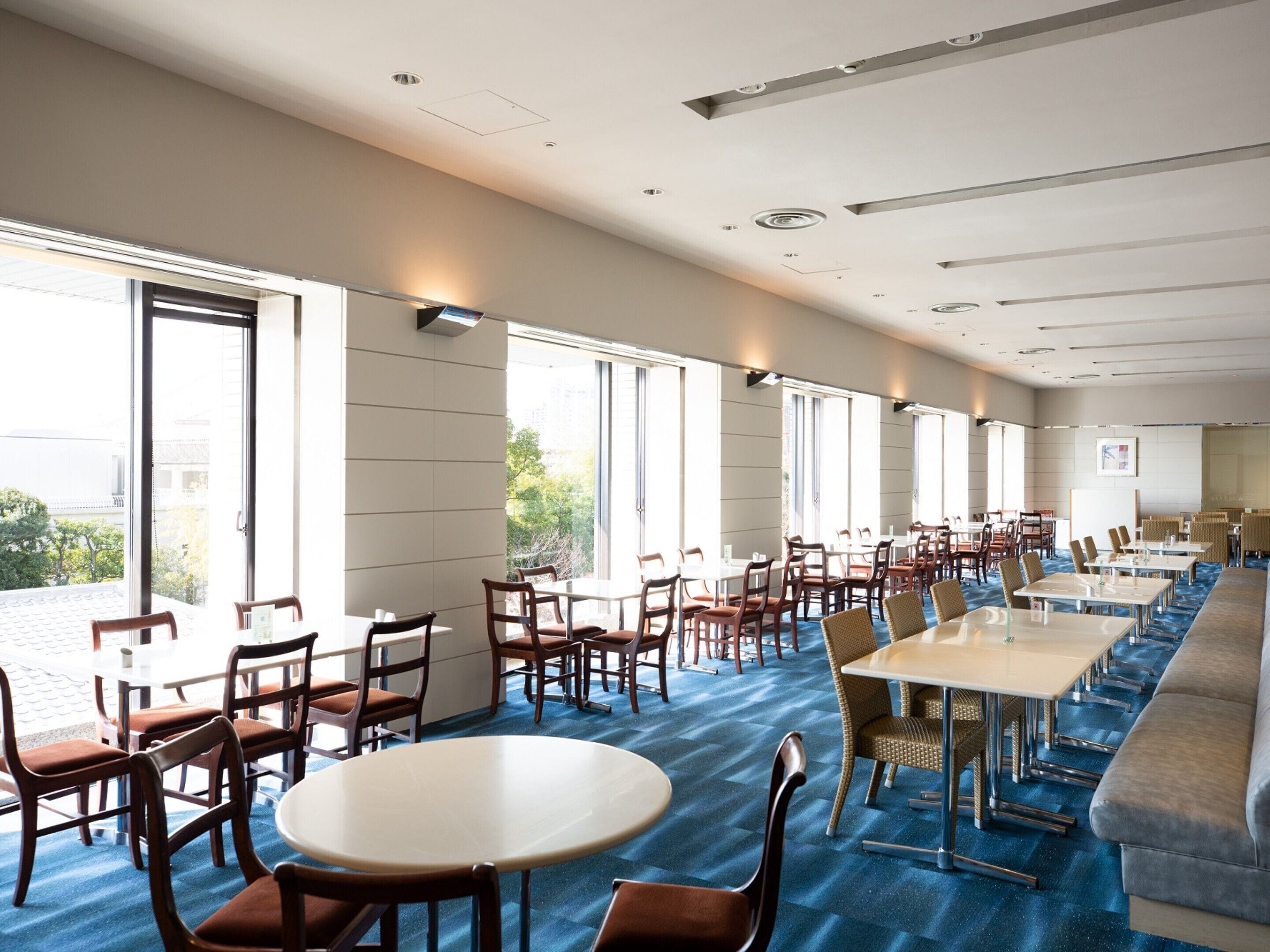 A spacious conference room with multiple tables and chairs, large windows, and a blue carpeted floor.