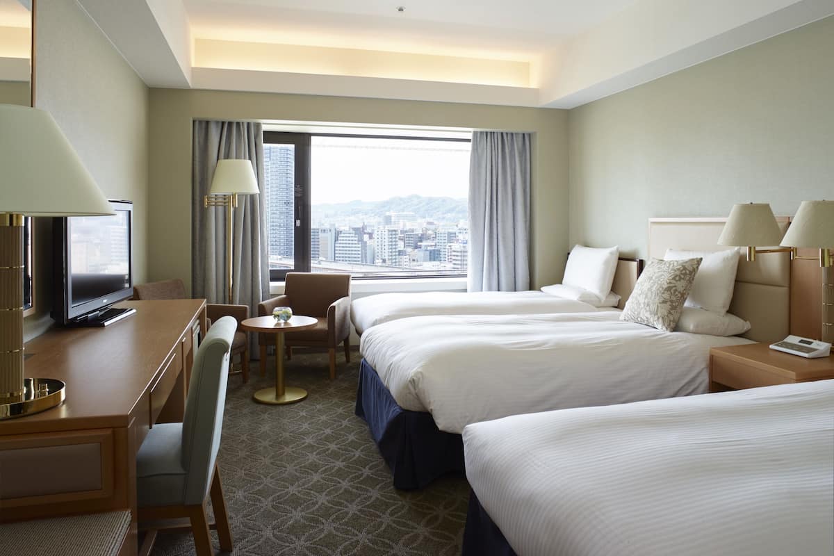 A hotel room with two beds, a desk with a TV, a chair, a small table, and a view of the city.