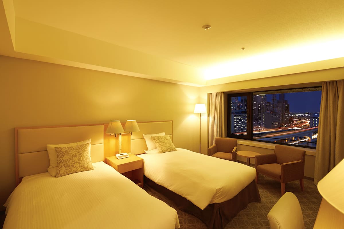 A hotel room with two beds, a desk, a chair, and a view of a cityscape at night.