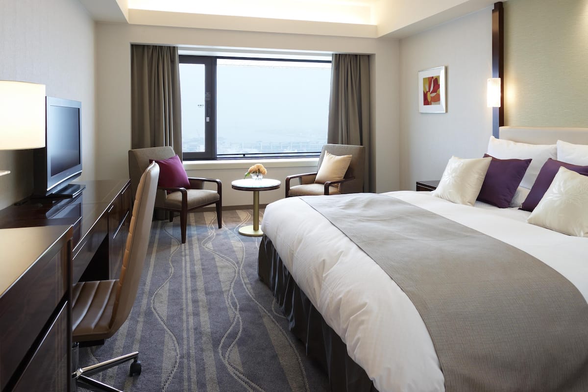 A hotel room with a large bed, a desk with a TV, two armchairs, a small round table with a vase, and a view of the city through a large window.