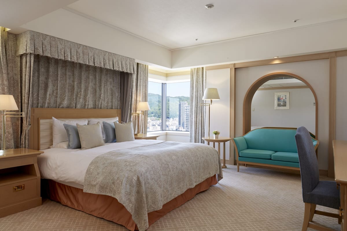 A hotel room with a large bed, a sitting area with a sofa, and a view of the city through large windows.