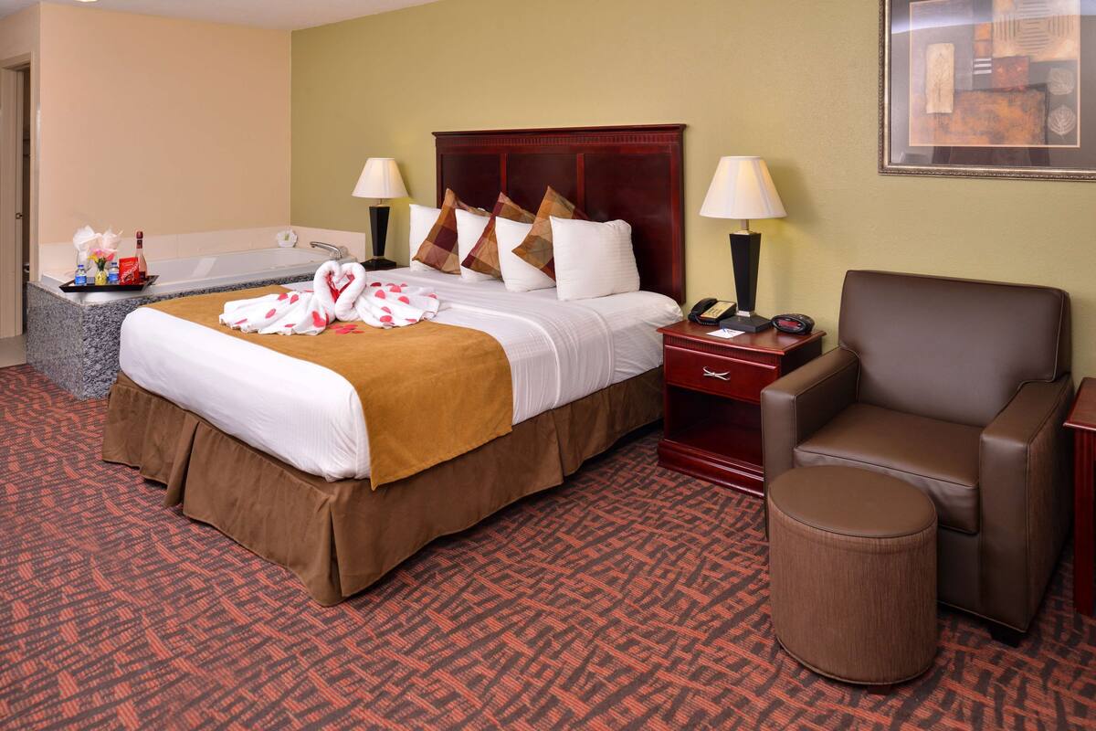 Une chambre d’hôtel avec un lit, deux tables de chevet, une chaise, un pouf rond, une baignoire et un tableau encadré au mur.