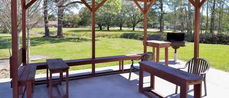 A covered outdoor seating area with picnic tables and chairs, a grill, and a view of a park with trees and grass.