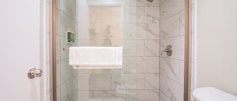 Une douche fermée par des parois de verre, avec un revêtement intérieur en carreaux de marbre, une serviette blanche accrochée à la porte et un pommeau de douche.