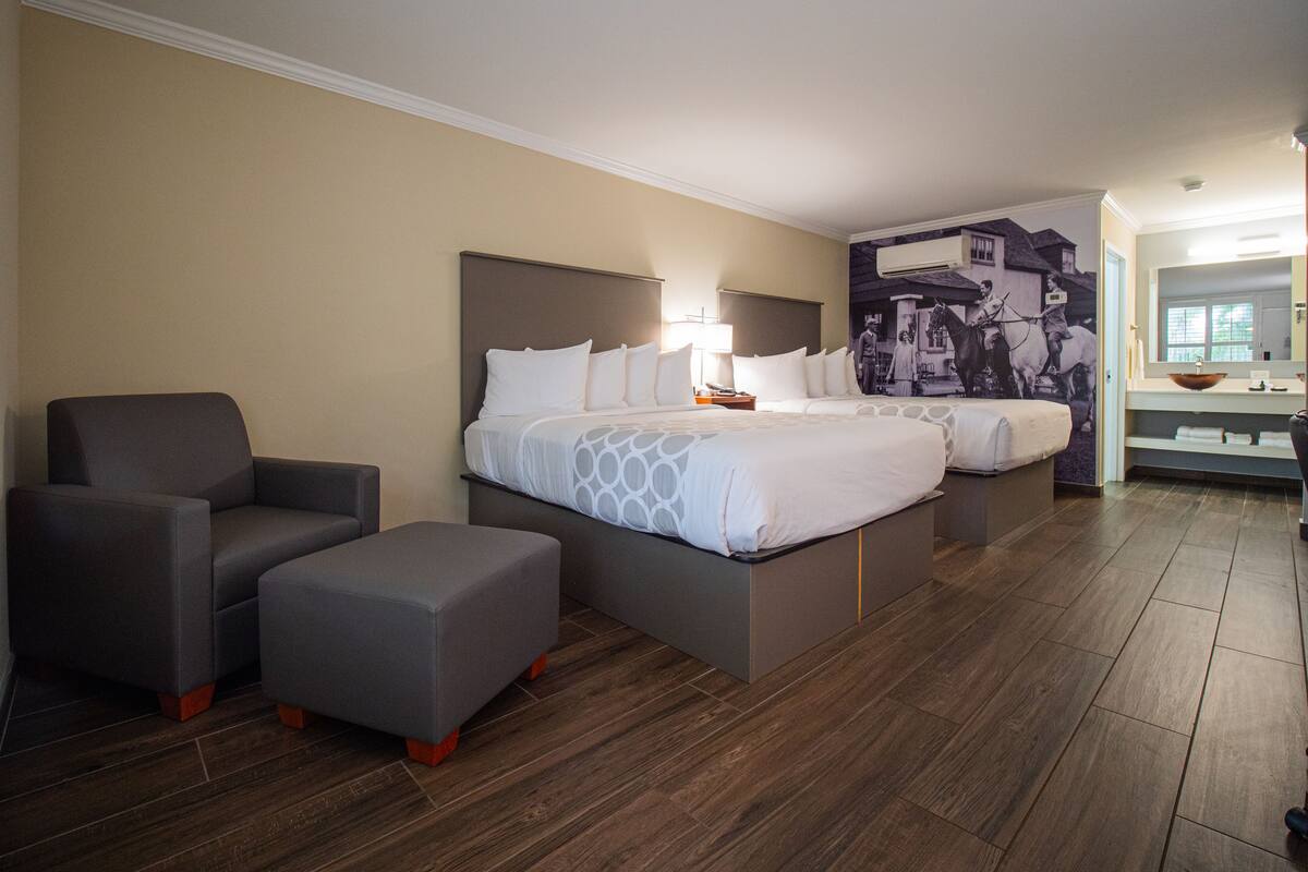 A hotel room with two beds, a chair, an ottoman, a nightstand, a wall-mounted air conditioner, and a bathroom with a sink and mirror.