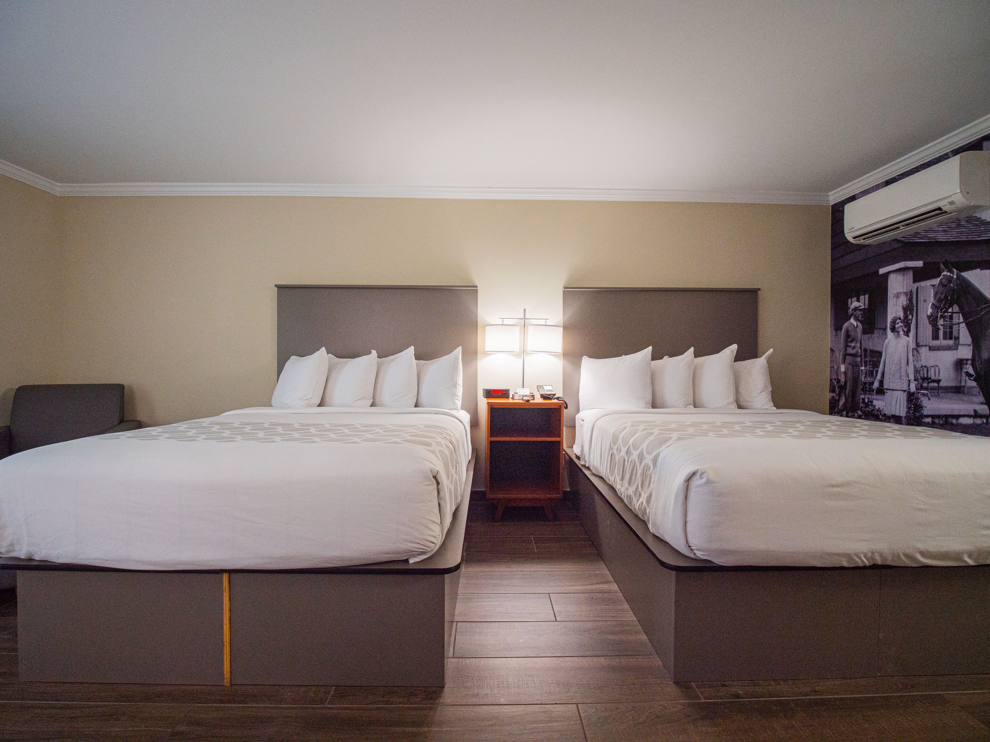 A hotel room with two single beds, a nightstand with a lamp, and a wall-mounted air conditioner.