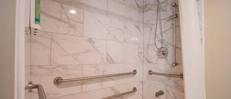 A bathroom with marble tiles, a bathtub, a shower with a handheld showerhead, and grab bars for accessibility.
