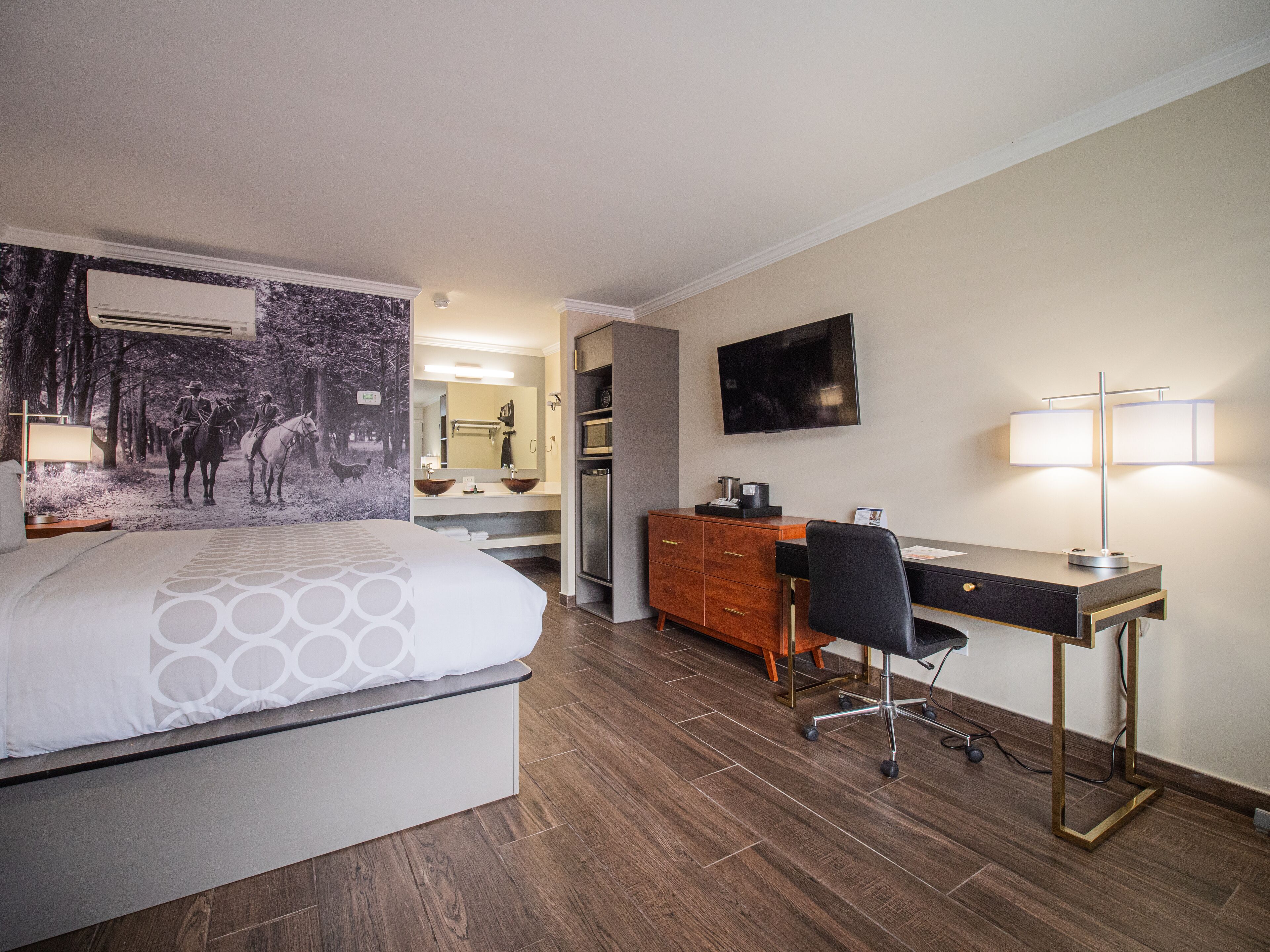 A hotel room with a bed, a desk, a chair, a TV, and a bathroom with a mirror and sink.