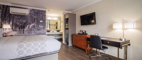 A hotel room with a bed, a desk, a chair, a TV, and a bathroom with a mirror and sink.