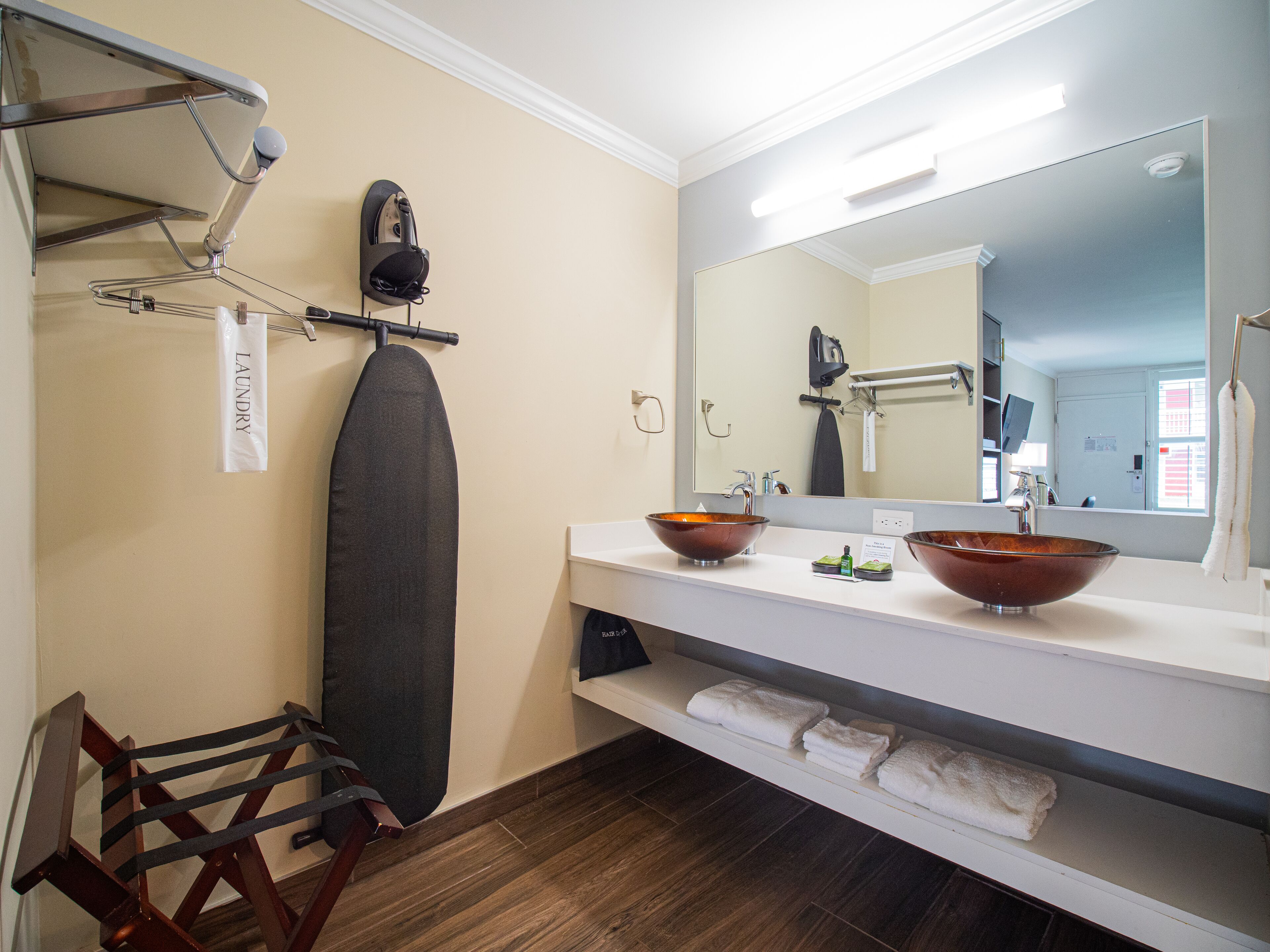 Un baño con un espejo grande, dos lavamanos con recipientes de cobre, un perchero para toallas y una silla de madera.