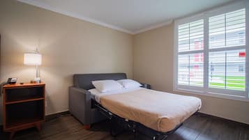 Una cama individual con cabecera gris, una mesita de noche con lámpara y una ventana con persianas.