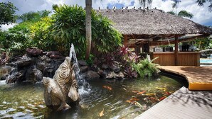Un bassin à carpes koï agrémenté d’une cascade et d’une sculpture de poissons, entouré de plantes tropicales et d’une structure à toit de chaume.
