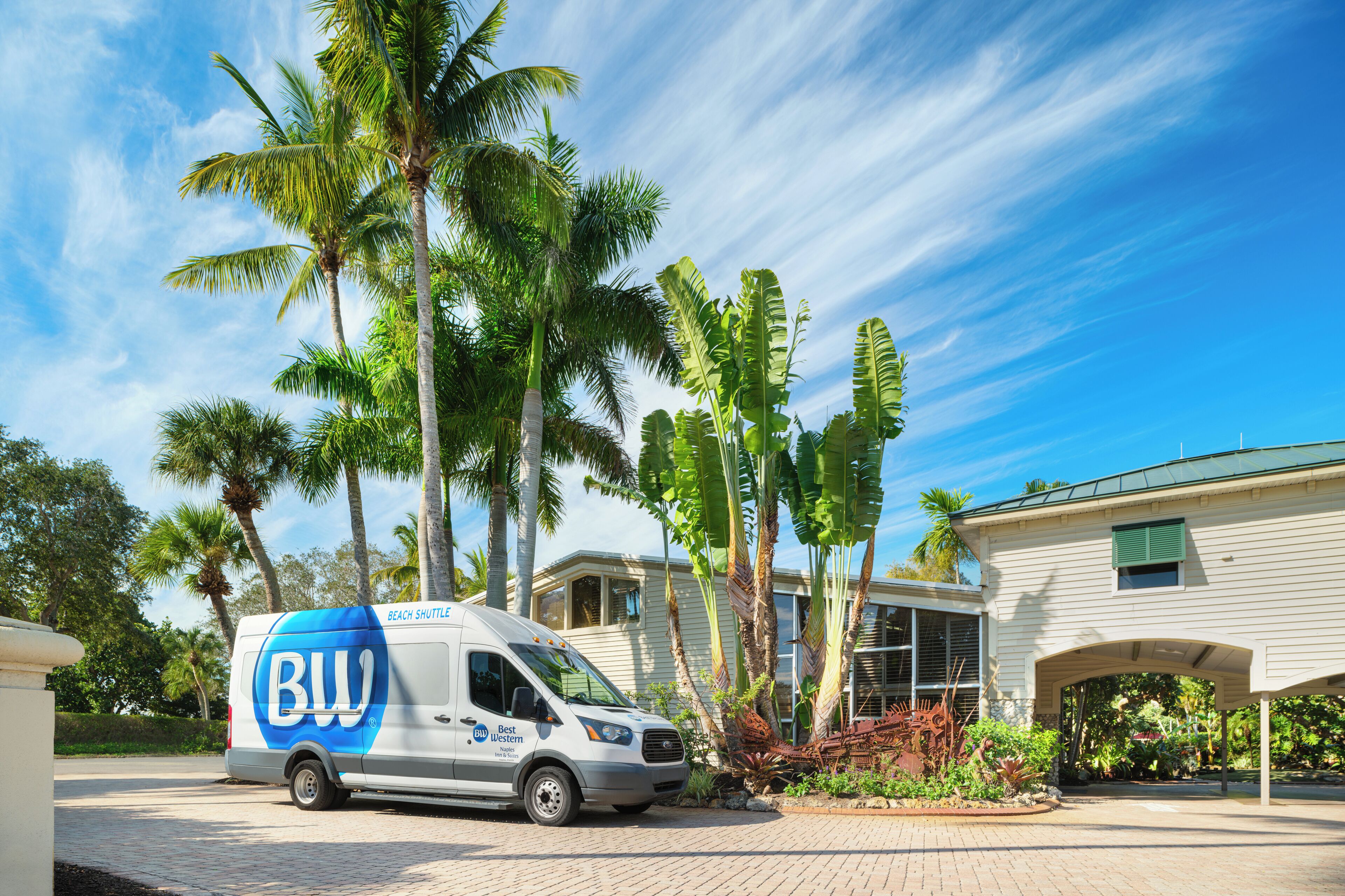 Une navette de plage arborant le logo « Best Western » est stationnée devant une maison bordée de palmiers.