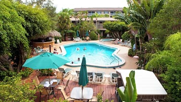 A pool area with lounge chairs and umbrellas, surrounded by lush greenery and a building in the background.