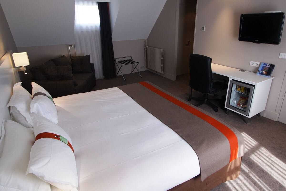 A hotel room with a large bed, a desk, a chair, a TV, and a mini-fridge.