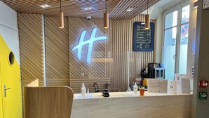 A modern reception area with a wooden slatted wall, a large illuminated 'H' sign, a coffee machine, and a menu board.