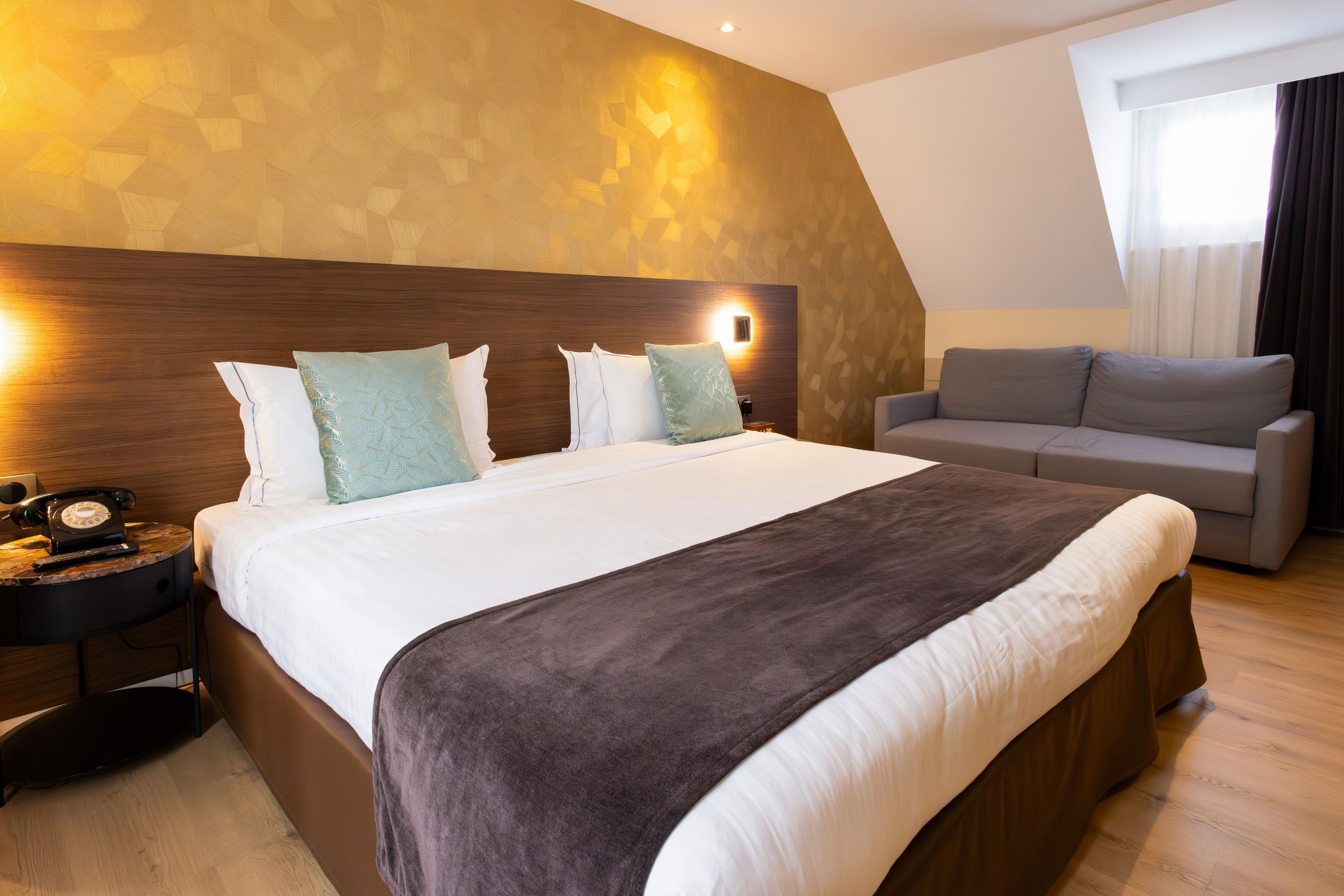 A hotel room with a large bed, a sofa, a bedside table with a telephone, and a wall-mounted light.