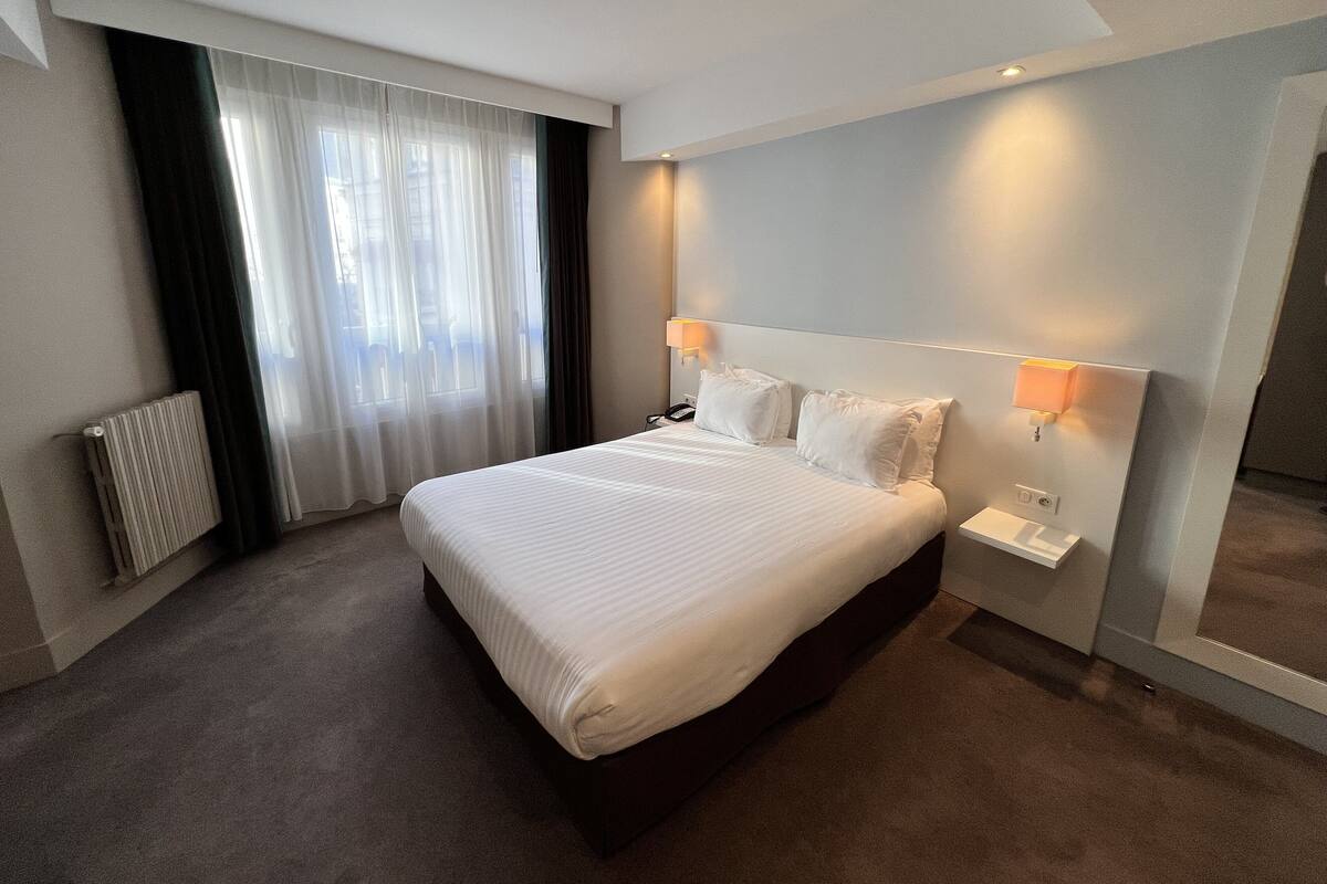 A hotel room with a large bed, a bedside table, a lamp, and a window with curtains.