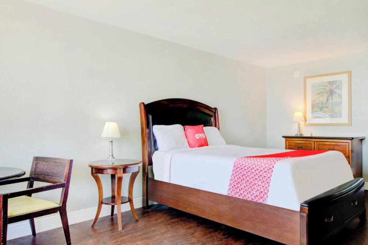 Une chambre d’hôtel avec un lit en bois, une petite table ronde, une chaise, une table de chevet et une lampe.